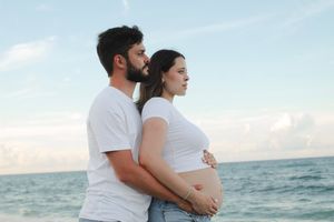 Man embracing pregnant woman from behind in white tops gazing at ocean horizon.