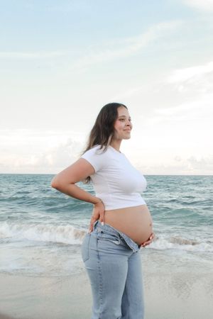 Smiling pregnant woman in white crop top and jeans cradling baby bump on beach.