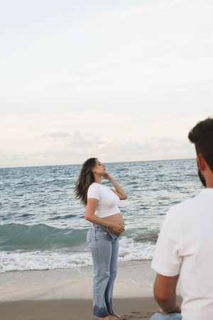 Pregnant woman in white top holding belly on beach while partner watches nearby.