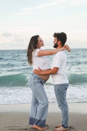 Pregnant couple embracing and smiling at each other on sandy beach with ocean waves.