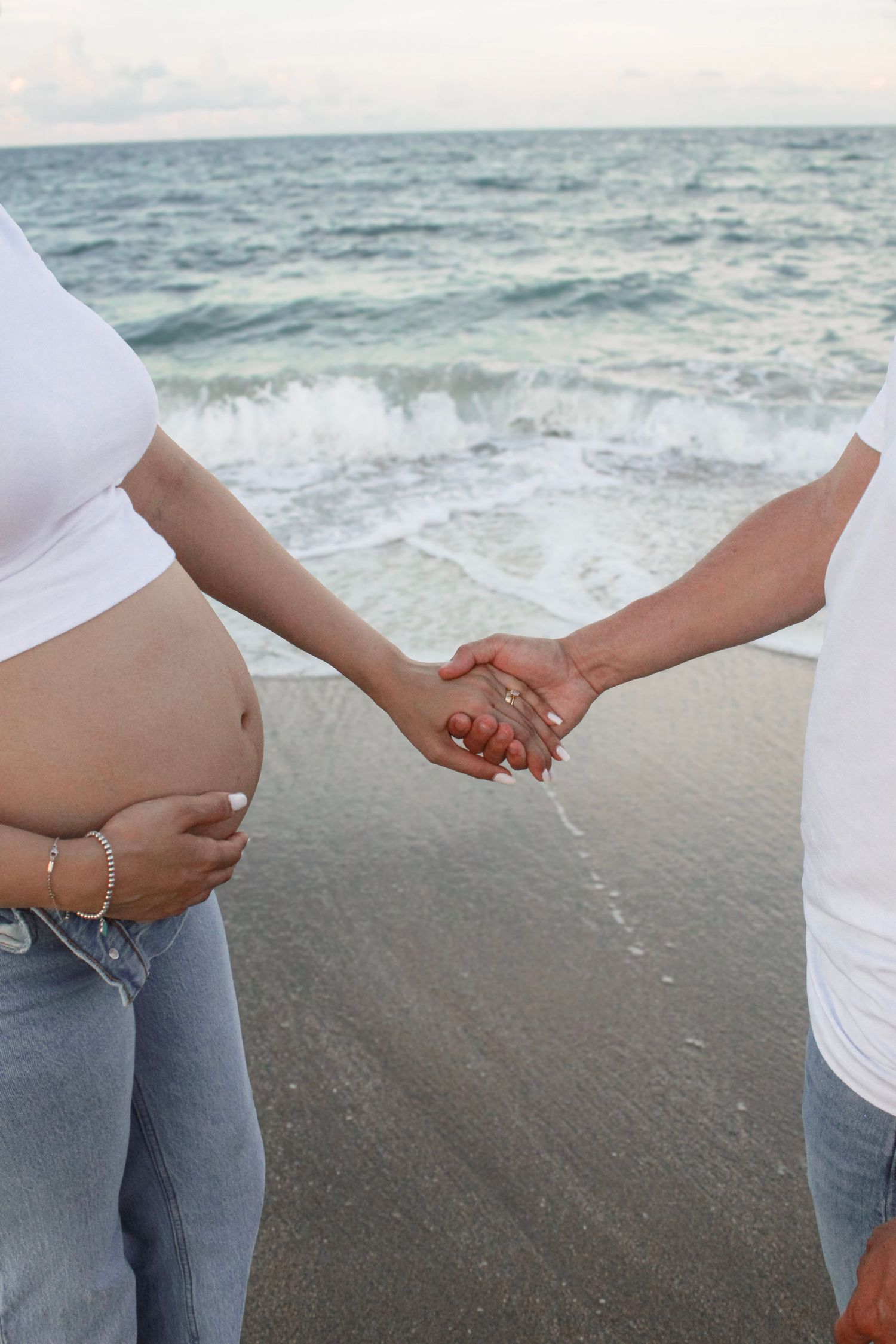 Pregnant couple holding hands on a sandy beach with ocean waves in the background.