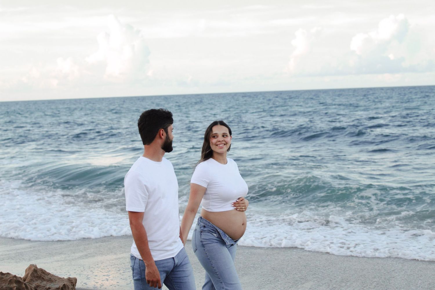 Pregnant couple in matching white tops and jeans walking on beach with ocean waves.