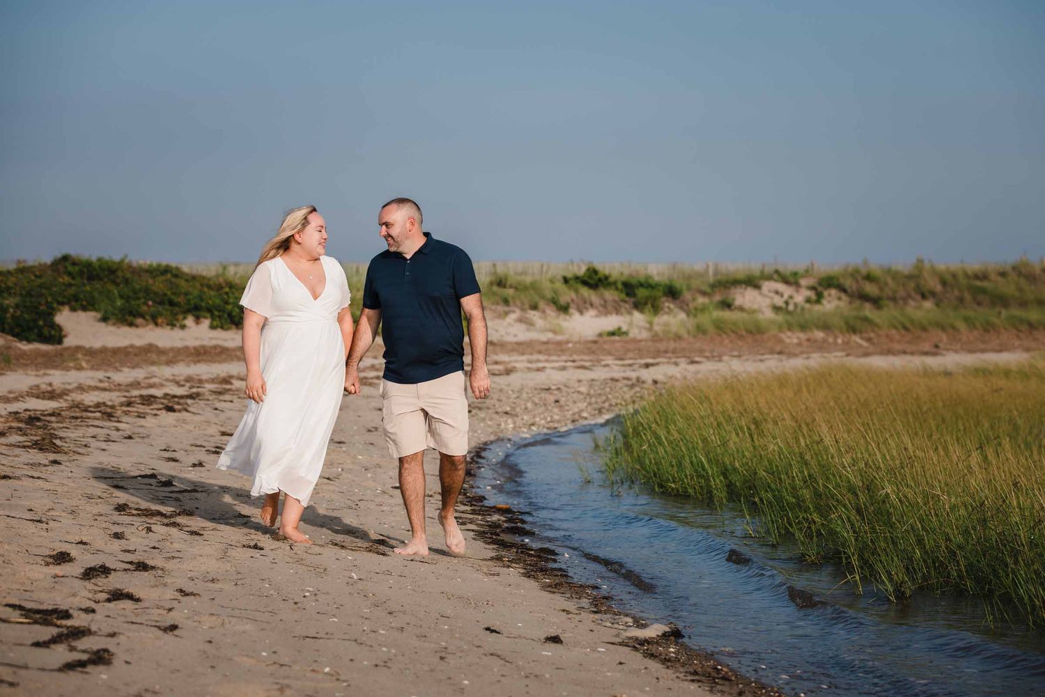Engaged couple holding hands and walking along the shoreline at Duxbury Beach, Massachusetts, in warm afternoon sunlight.