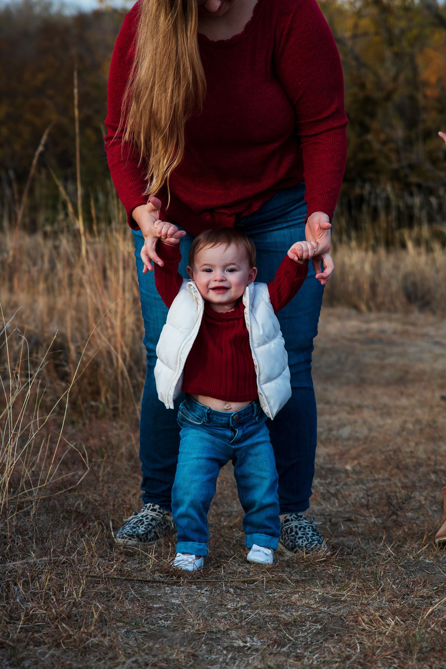 A mother helps her smiling baby take first steps on a rural path, both wearing matching red sweaters and jeans in autumn.