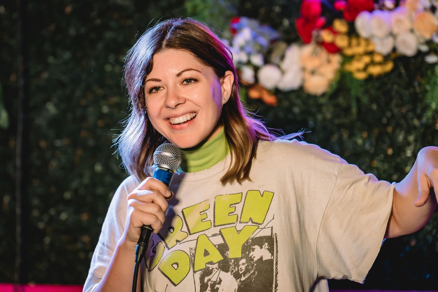 Jessie "Jetski" Johnson comedian Woman in Green Day t-shirt performing stand-up comedy on stage holding microphone with floral backdrop.