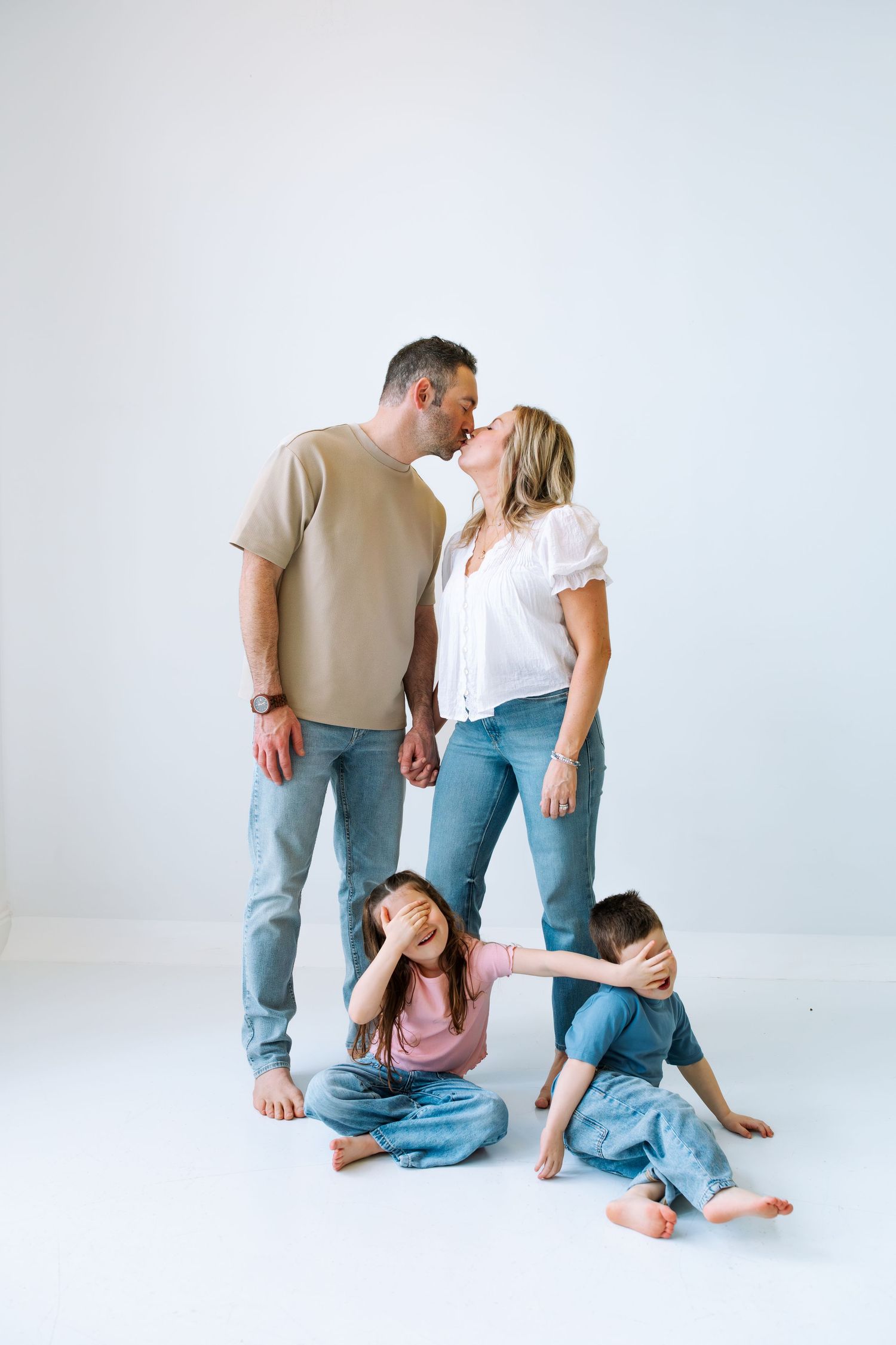 A family photo with parents kissing while two young children sit on the floor covering their eyes.