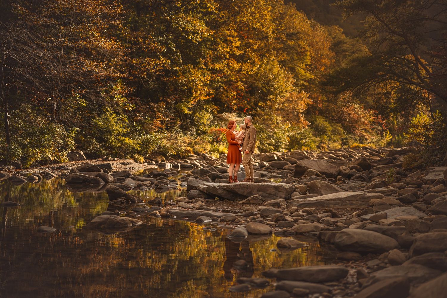 Person in red dress standing on rocks by a reflective stream surrounded by vibrant autumn foliage.