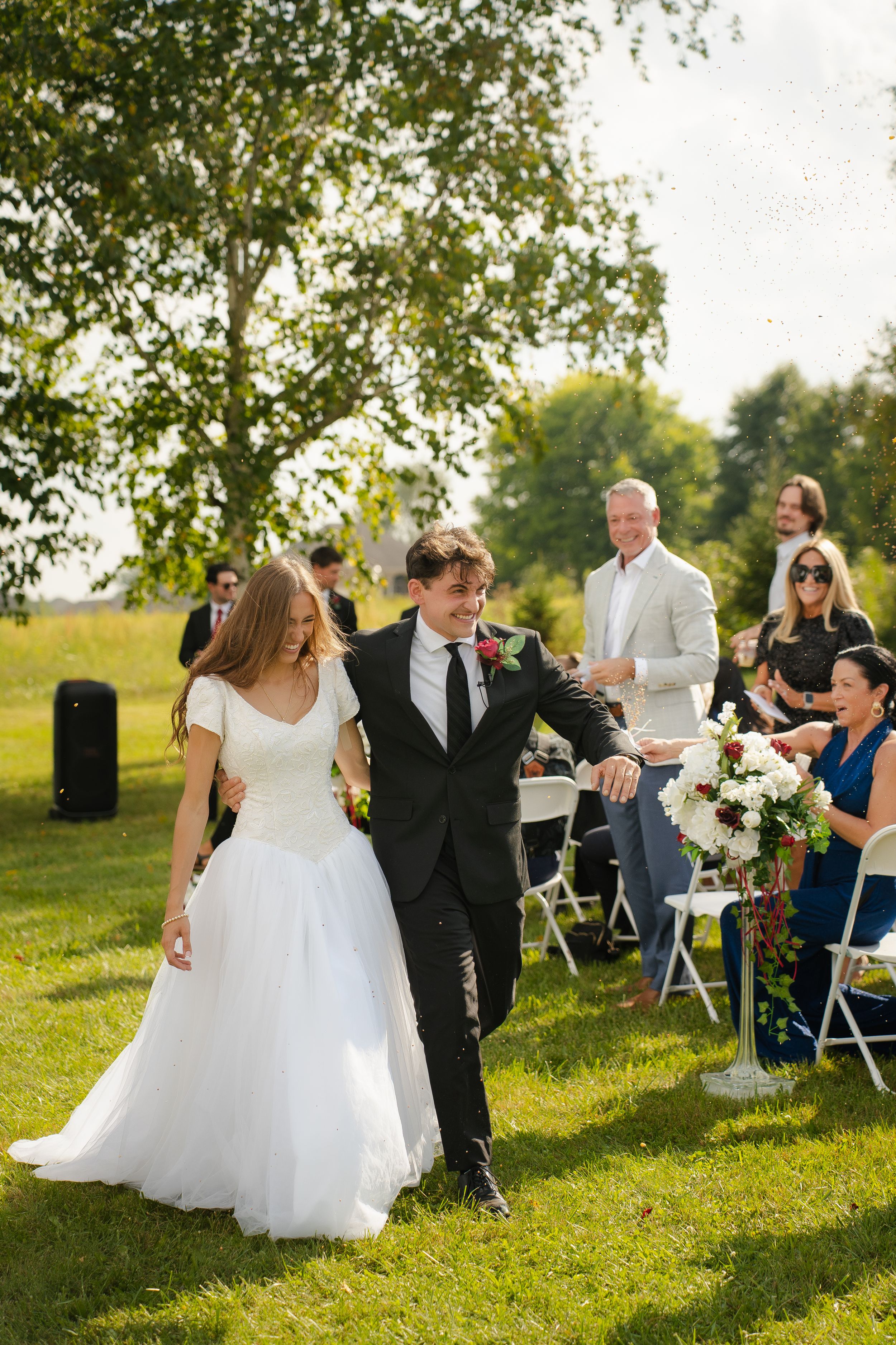 Bride and groom walk down the aisle at an outdoor wedding ceremony on a sunny green lawn with guests celebrating.