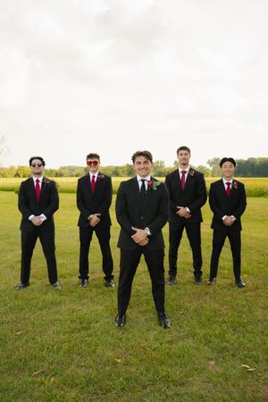 Five groomsmen in black suits and red ties posing outdoors on green grass for a wedding photo.