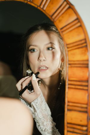 Young woman applying lipstick while looking in an ornate wooden framed mirror, wearing lace sleeves and pearl earrings.