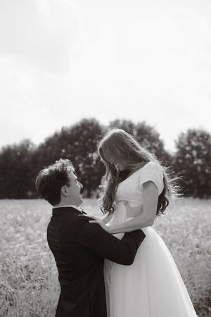 Black and white photo of a bride and groom embracing in a field, gazing at each other romantically.