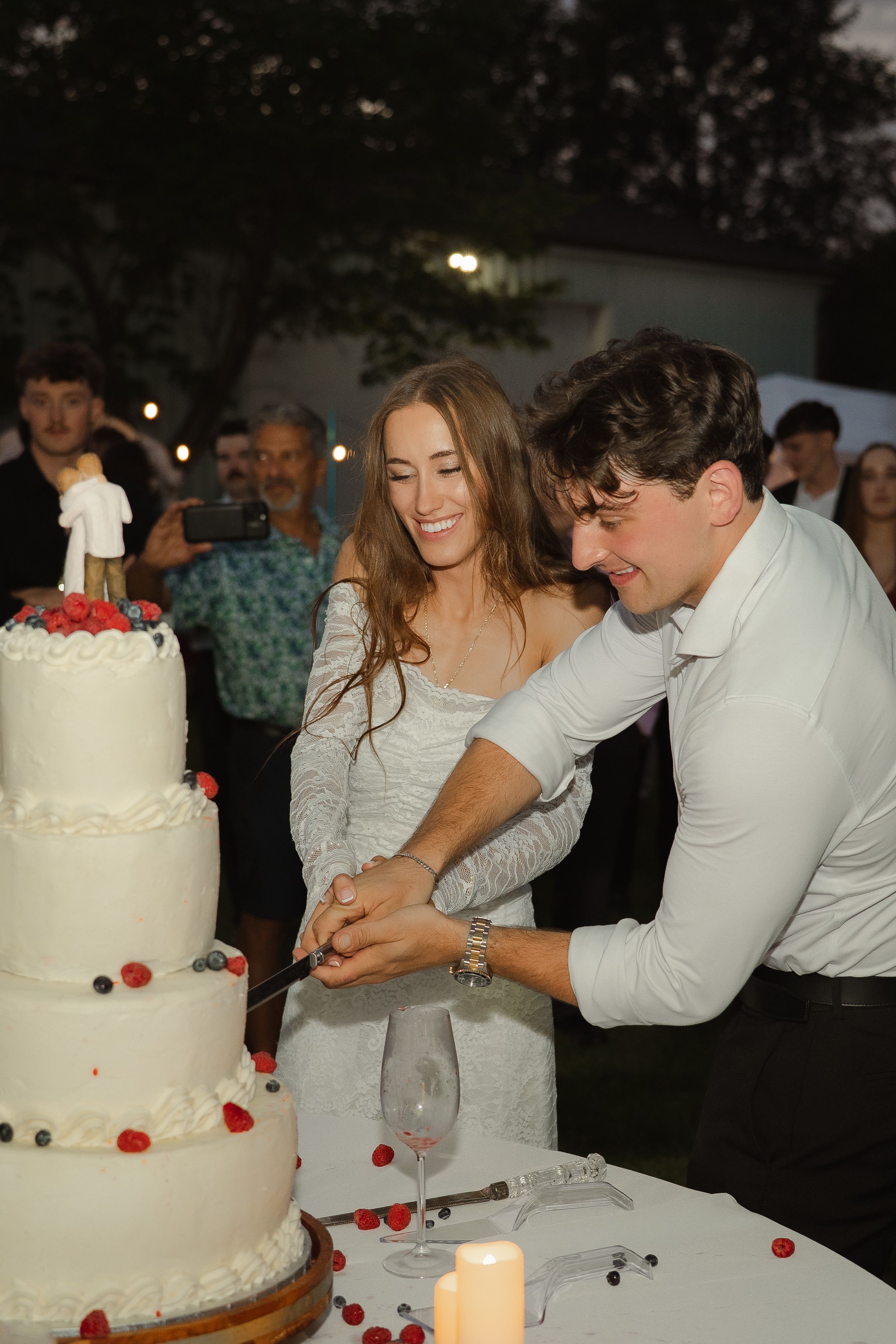 Bride and groom cutting a white tiered wedding cake decorated with fresh berries at an outdoor evening reception.