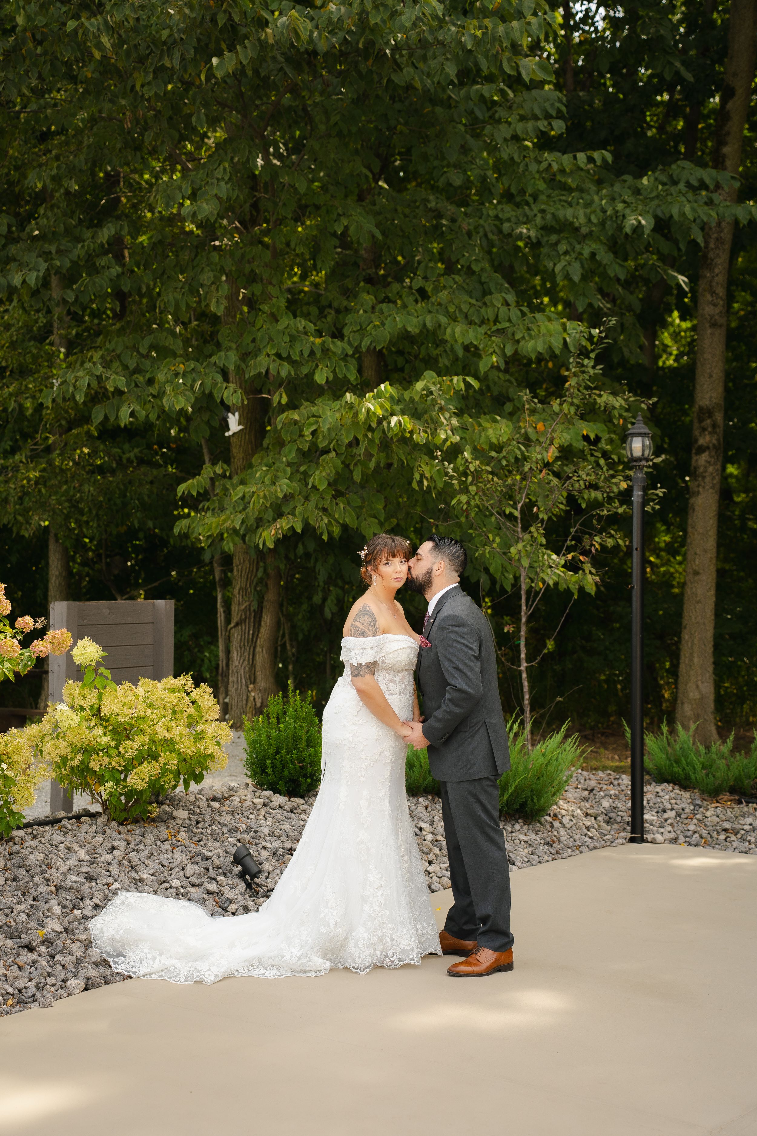Bride and groom share a romantic kiss outdoors on their wedding day, surrounded by lush green trees and landscaped gardens.