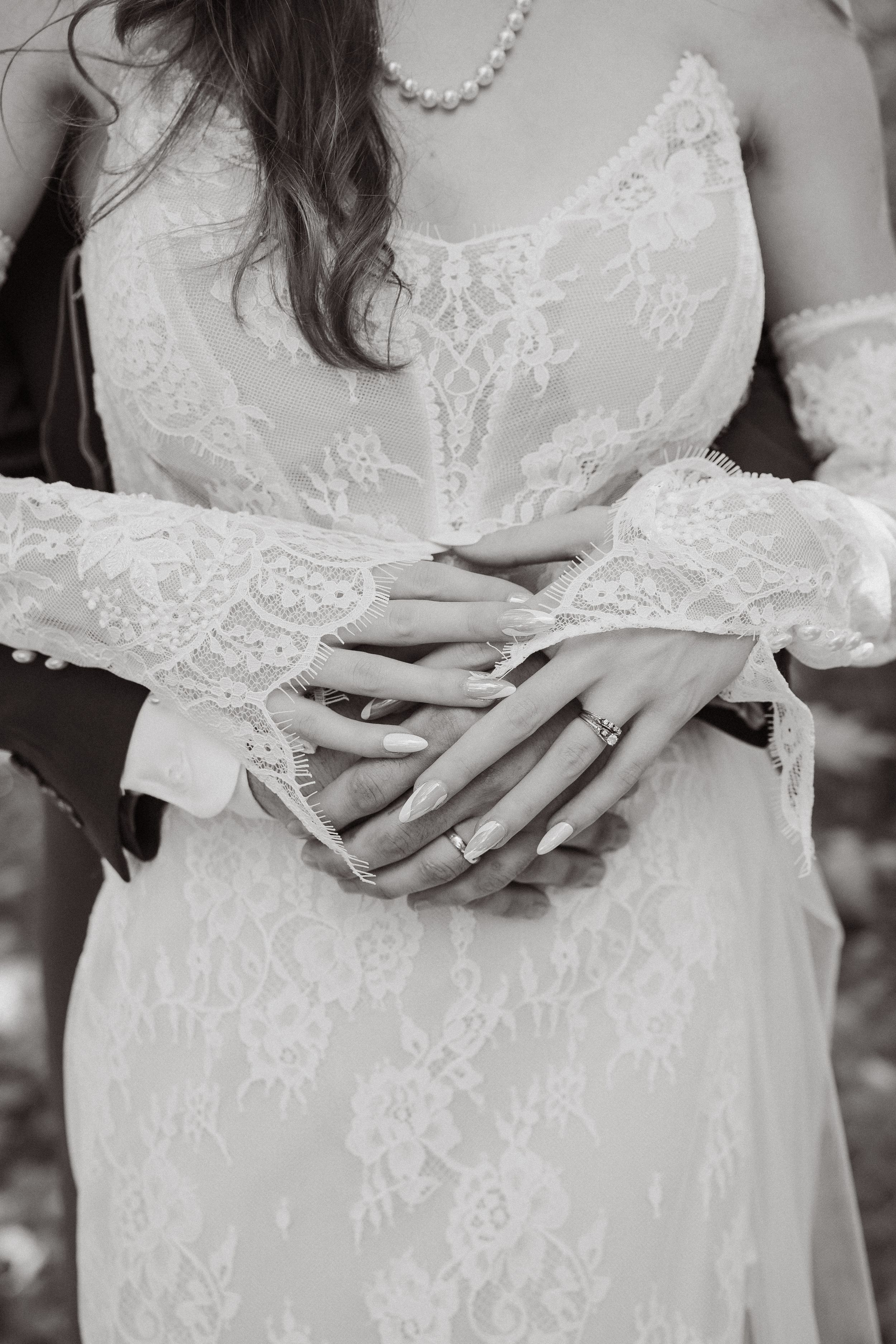 Black and white photo of bride and groom hands intertwined, showcasing wedding ring on lace dress.