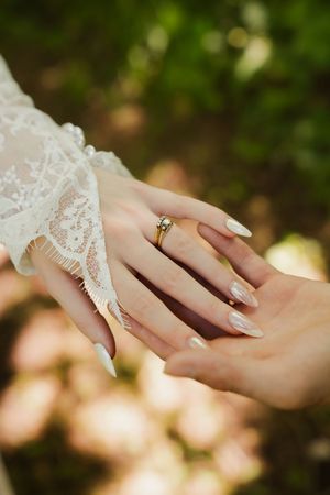 Close-up of bride's hands wearing a gold engagement ring with elegant white almond-shaped nails and lace wedding dress sleeve.