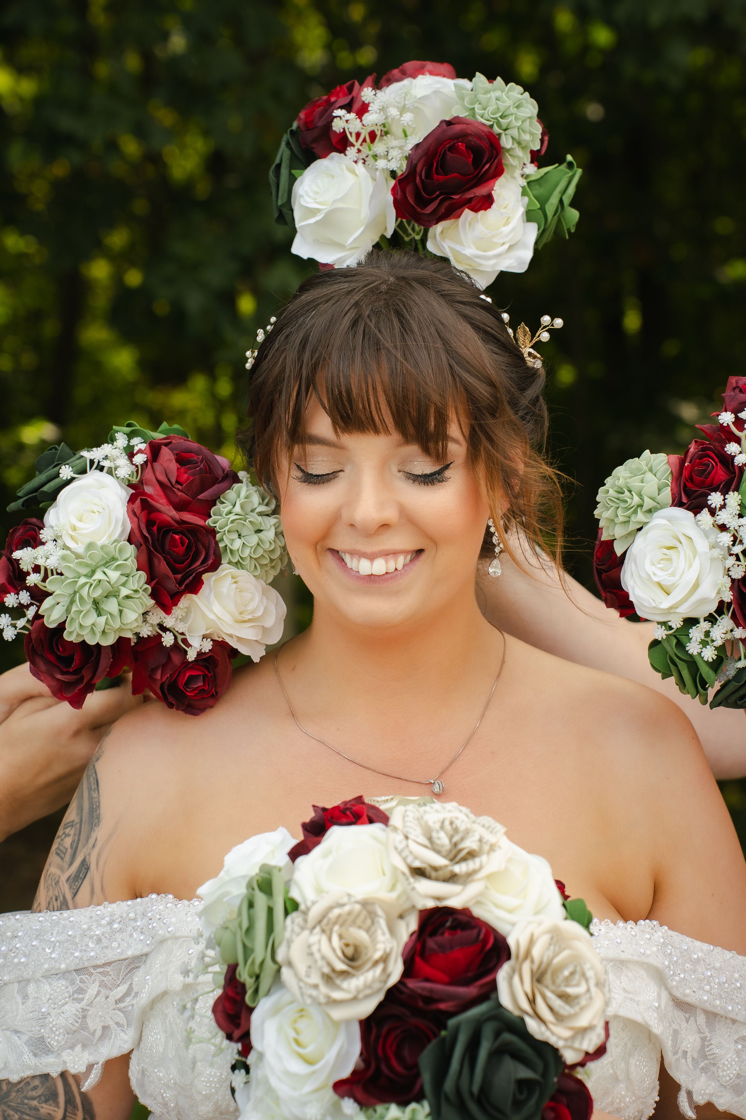 Smiling bride with eyes closed holding red and white rose bouquet, wearing lace off-shoulder gown and floral hair arrangement.
