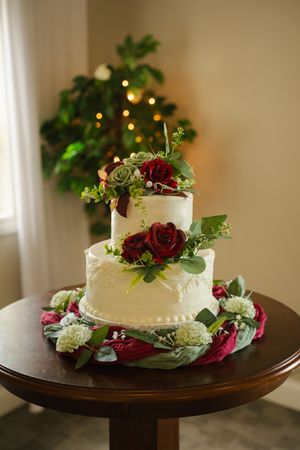 Elegant two-tier white wedding cake adorned with red roses and greenery on a wooden table with floral wreath base.