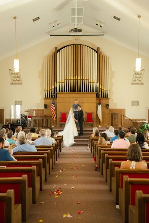 Wedding ceremony in a church with bride and groom at the altar, flower petals on the aisle, and guests seated in wooden pews.
