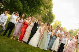 Large group of elegantly dressed teens posing outdoors at prom, wearing formal gowns and suits on a sunny day.
