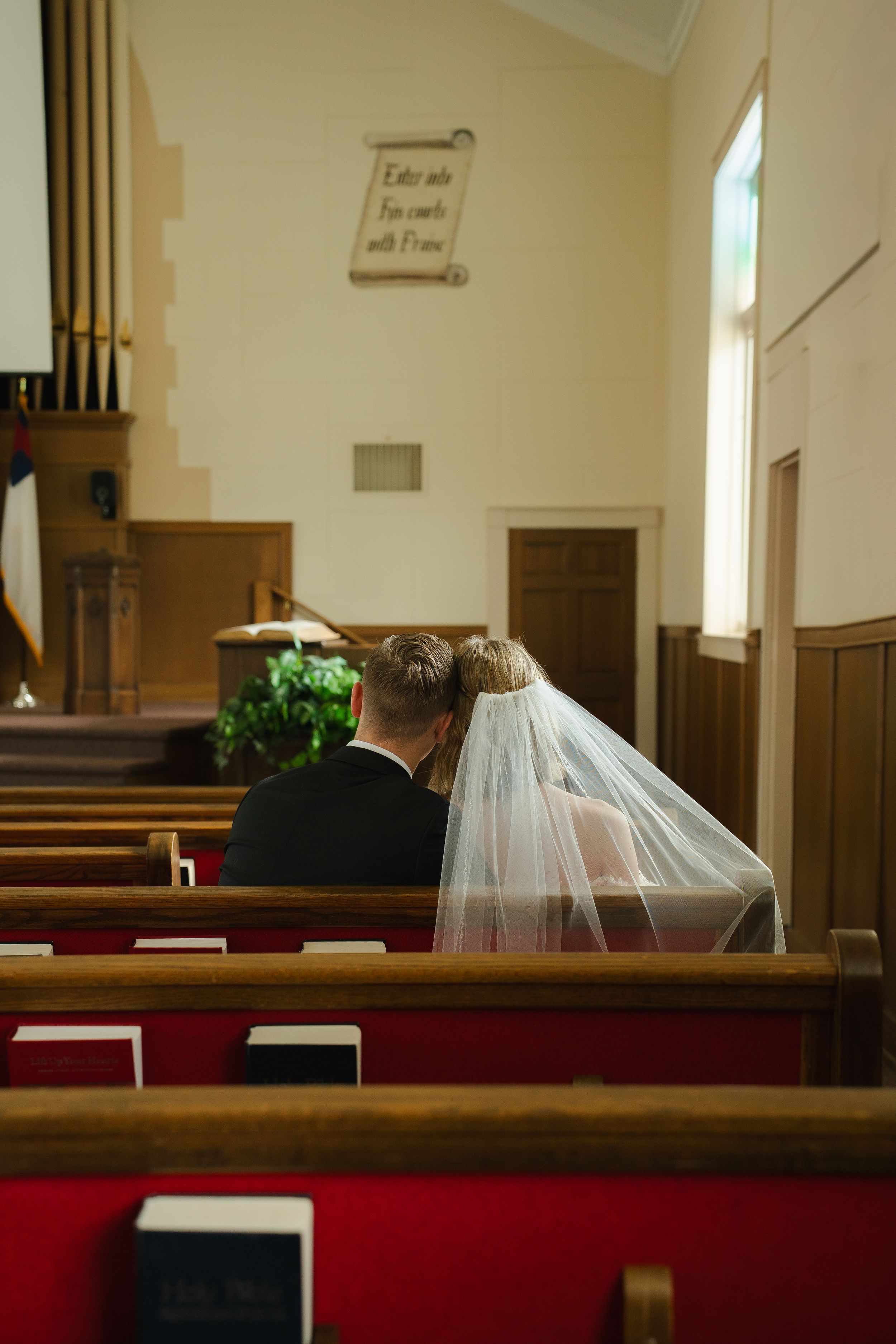 Bride and groom sitting together in wooden church pews during wedding ceremony, bride wearing white veil.