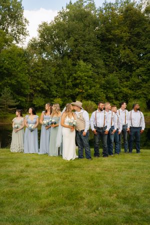 Wedding party poses outdoors on green lawn, bridesmaids in blue gowns, groomsmen in suspenders, couple kissing in center.