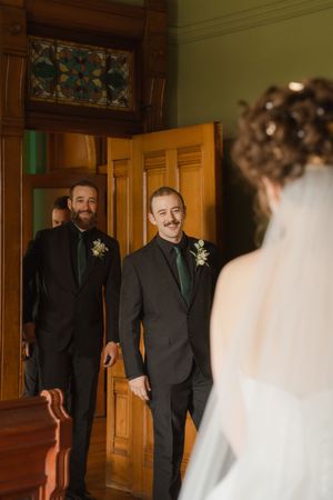 Groom smiling warmly at bride during first look moment inside elegant wooden doorway at wedding venue.