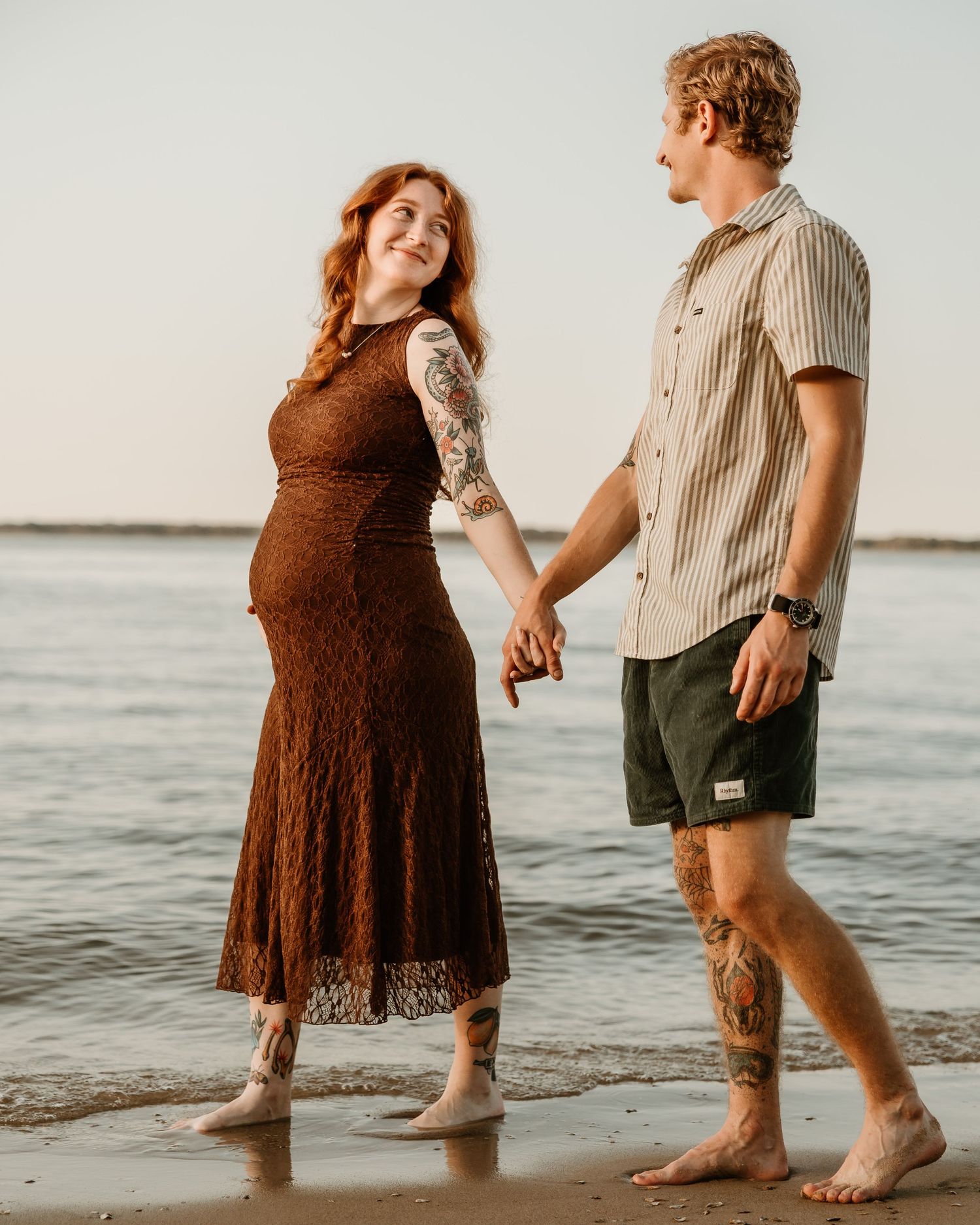Pregnant woman in brown lace dress holds hands with tattooed man in striped shirt walking barefoot on beach at sunset.