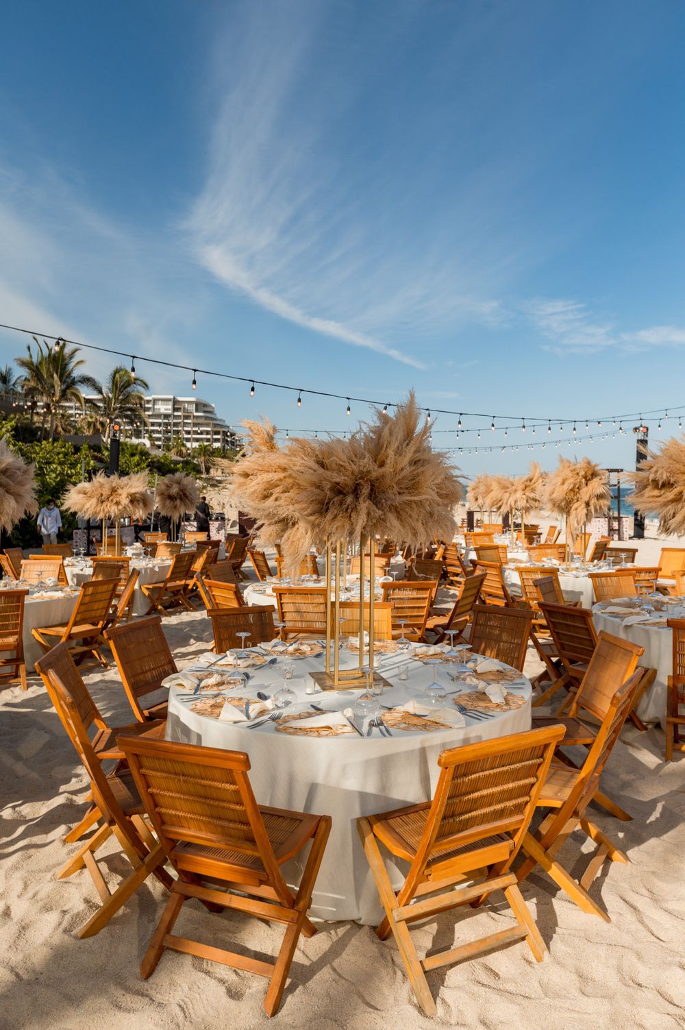 Elegant beach wedding reception with wooden chairs, round tables, pampas grass centerpieces, and string lights under a blue sky.
