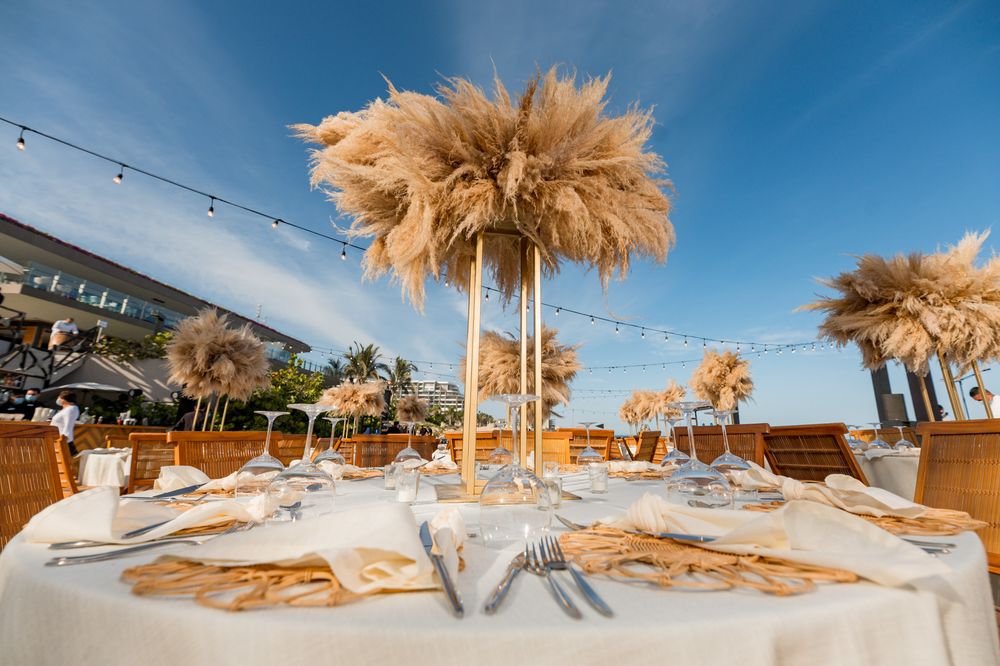 Elegant outdoor wedding reception table with tall pampas grass centerpieces, gold accents, and string lights by the ocean.