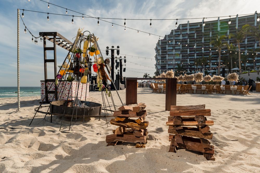 Beach setup with wooden pallets, decorative structures, and string lights near a modern hotel on a sandy shore.