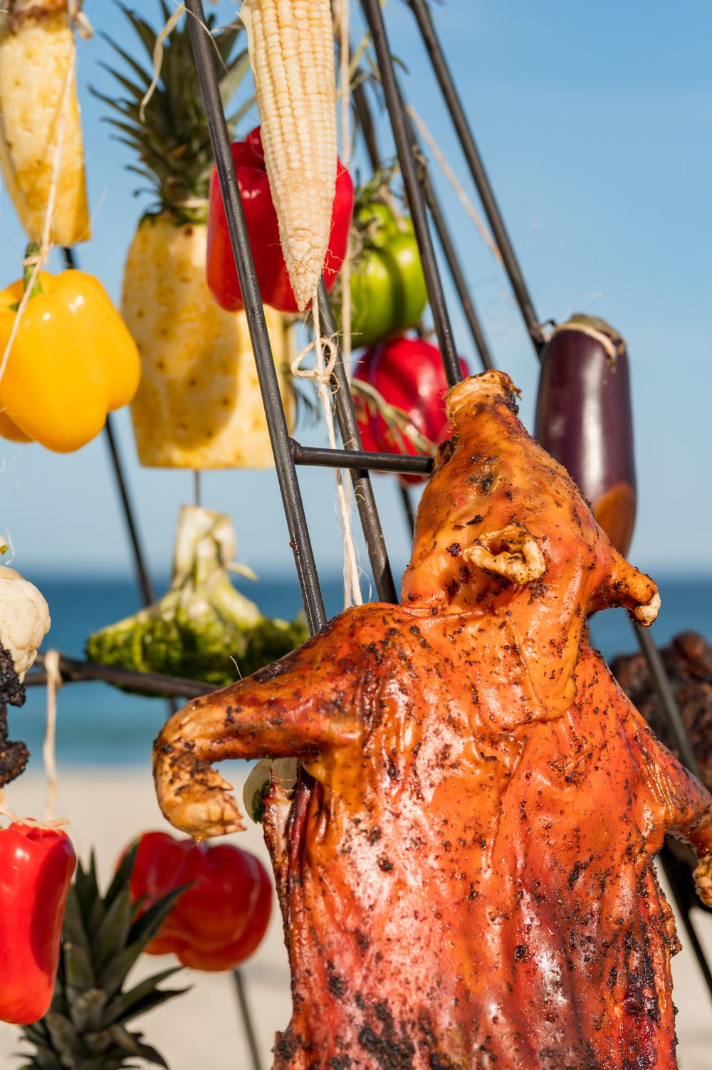 Roasted whole pig and colorful vegetables hanging on display hooks at a beachside outdoor cooking station.