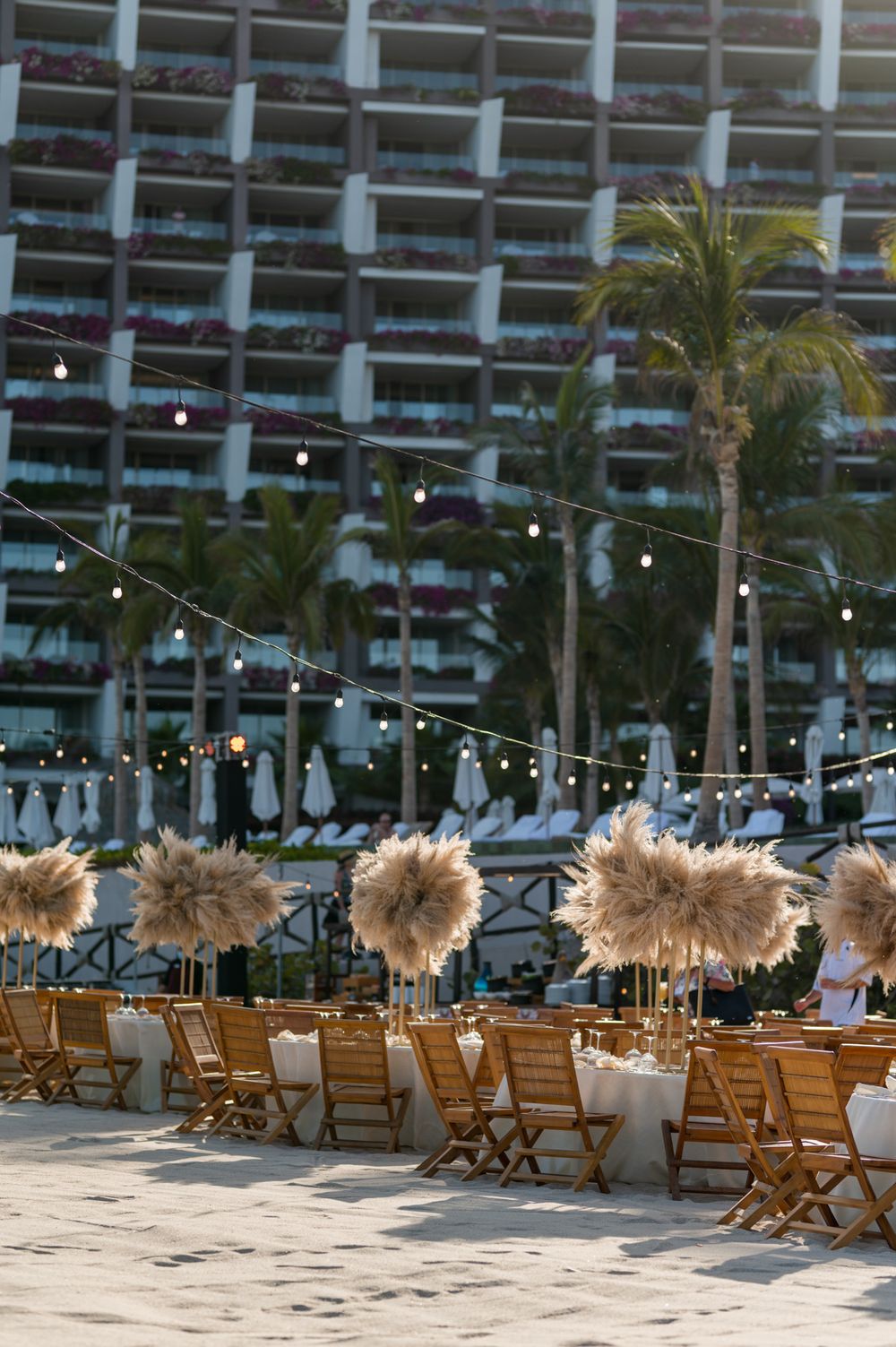 Beach wedding reception setup with wooden chairs, pampas grass centerpieces, string lights, and a resort backdrop at dusk.