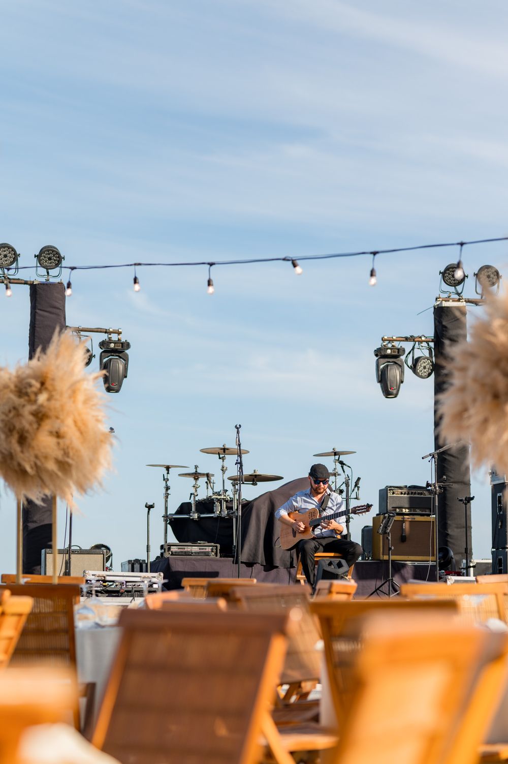 Guitarist performs on an outdoor stage with string lights, speakers, and drum kit under a blue sky.