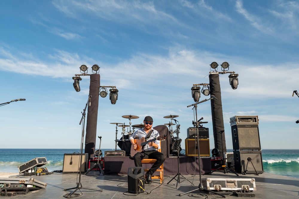 Musician plays guitar on outdoor beach stage with speakers, drums, and ocean backdrop under blue sky.