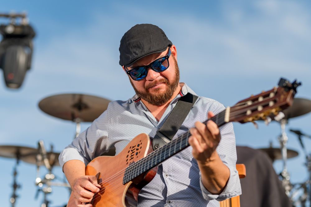 Bearded male guitarist wearing sunglasses and cap playing acoustic guitar outdoors at a live music event.