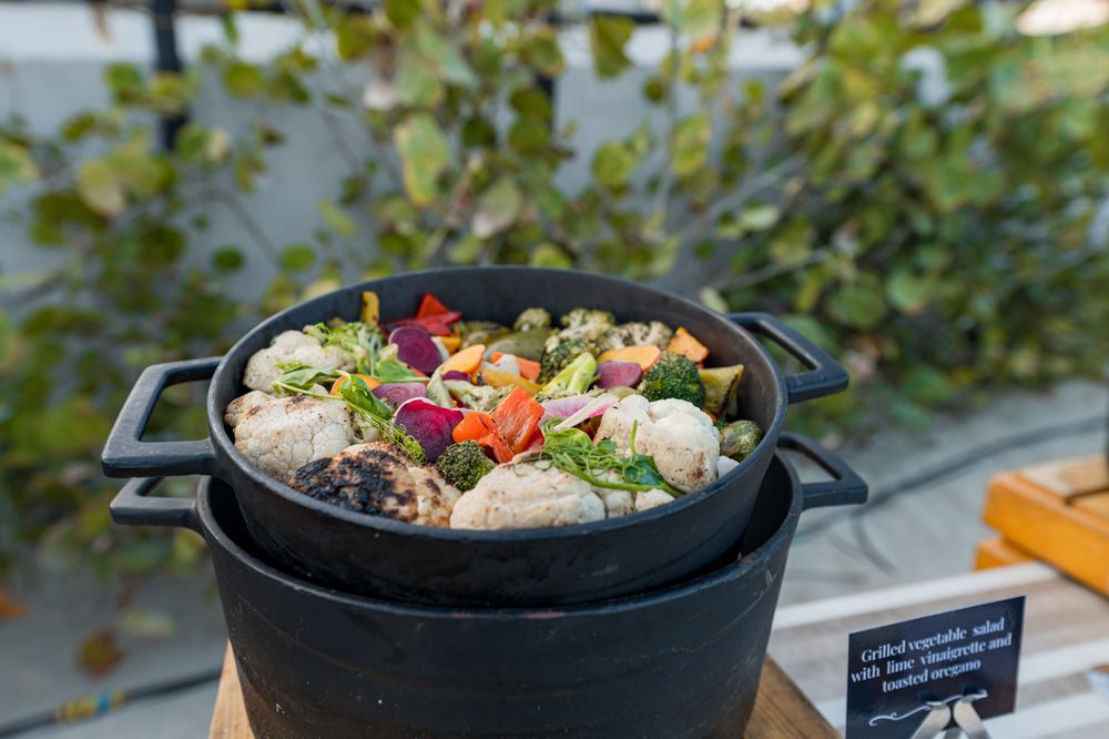 Black cast iron pot filled with grilled vegetables, meatballs, and edible flowers at an outdoor buffet.