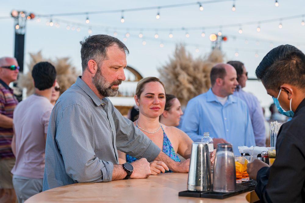 Couple at outdoor bar with string lights at dusk, conversing with masked bartender at a social gathering.