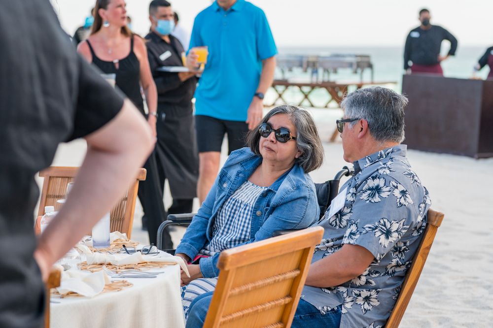 Two people in sunglasses chat at an outdoor beachside table during a casual daytime event.