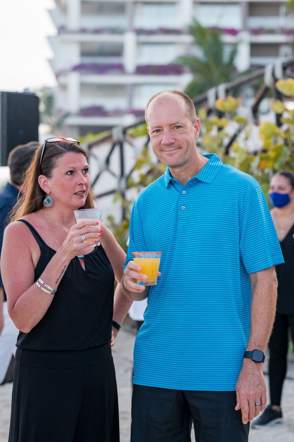 A man in a blue polo and woman in black dress hold tropical drinks at an outdoor beachside event.