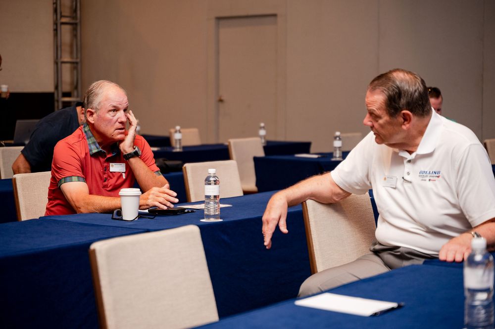 Two men in casual shirts engaged in conversation at a conference table with water bottles in a meeting room.