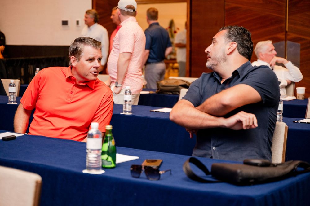Two men in casual attire engage in conversation at a blue-draped conference table during a business networking event.