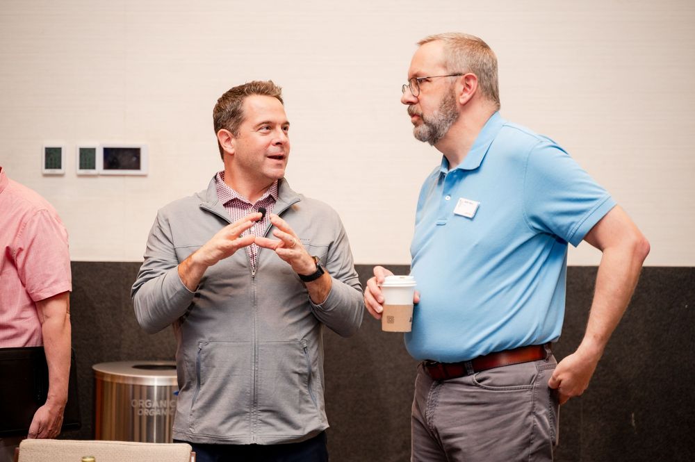 Two men engaged in conversation at a casual indoor networking event, one gesturing with hands while wearing a gray shirt.