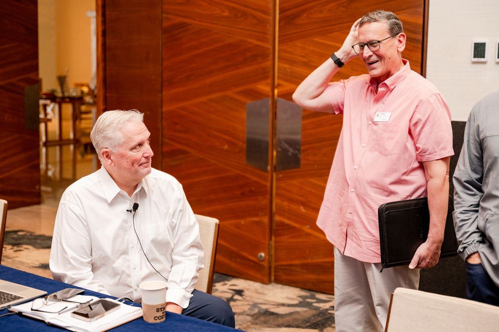 Two men in casual attire conversing at a conference table, one seated with coffee, the other standing holding a laptop.