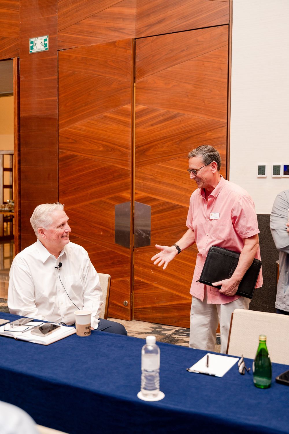 Two men in business casual attire converse at a conference table with a blue tablecloth in a modern venue.