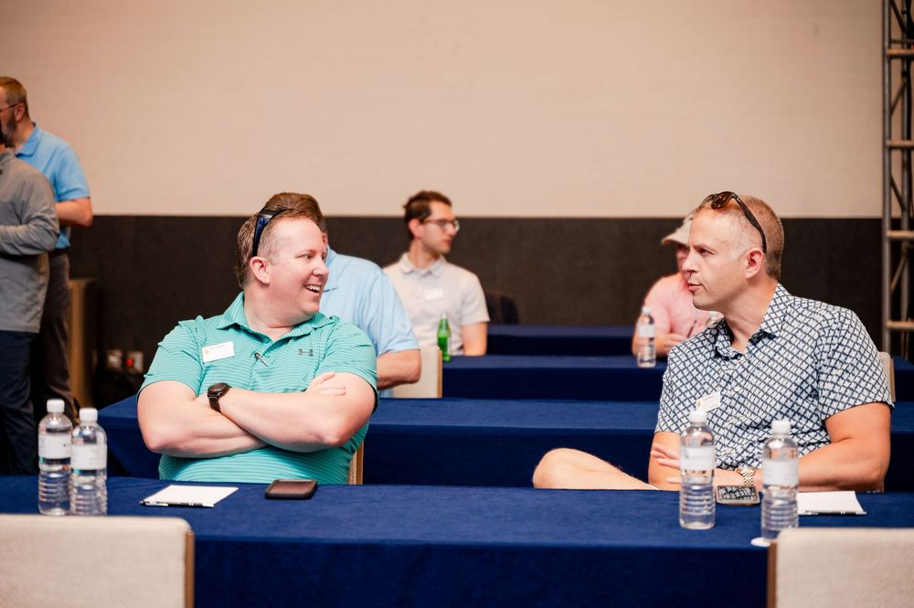 Two men sit and chat at a conference table in a modern auditorium setting with other attendees in the background.