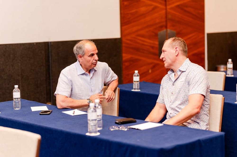 Two men sit at a blue-draped table in conversation during a professional meeting or conference event.