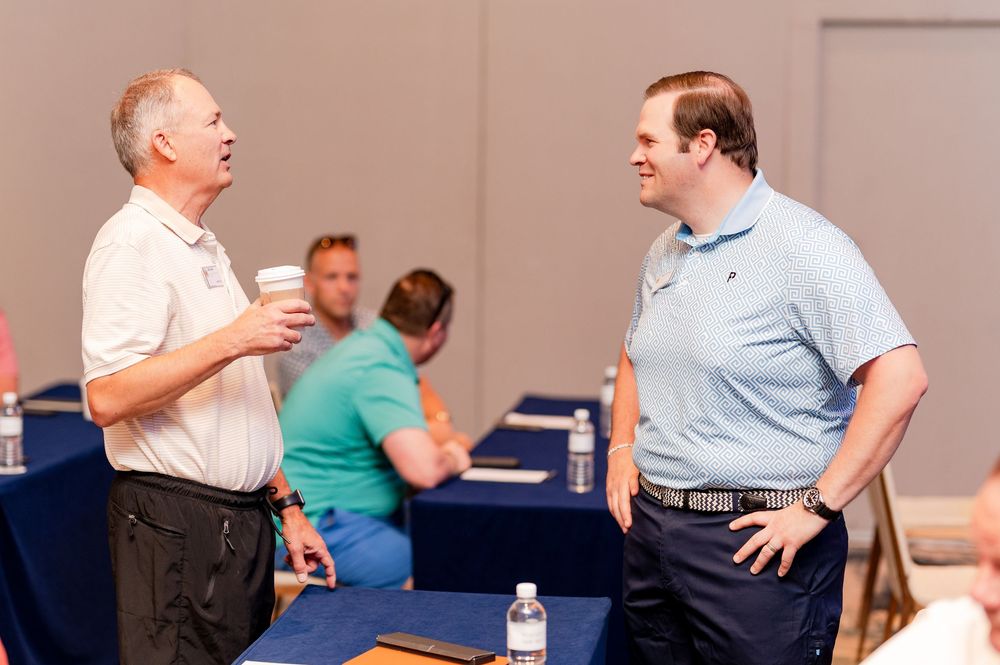 Two men in polo shirts engage in conversation at a professional networking event in a conference room.