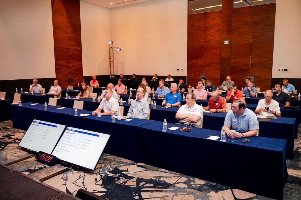 Professionals attending a corporate conference seminar seated at blue-draped tables in a modern hotel ballroom.