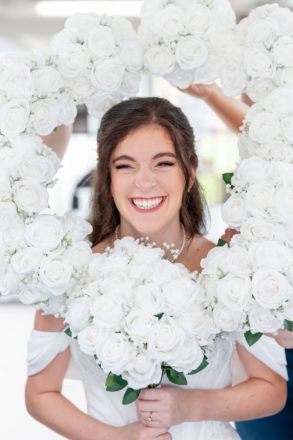 Smiling bride in white dress holds bouquet of white roses framed by a heart-shaped floral arrangement.
