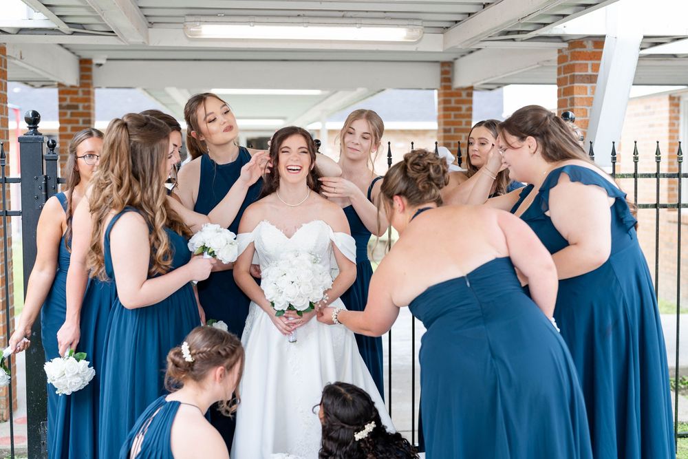 Bride in white gown holds bouquet surrounded by bridesmaids in navy blue dresses during wedding preparations.